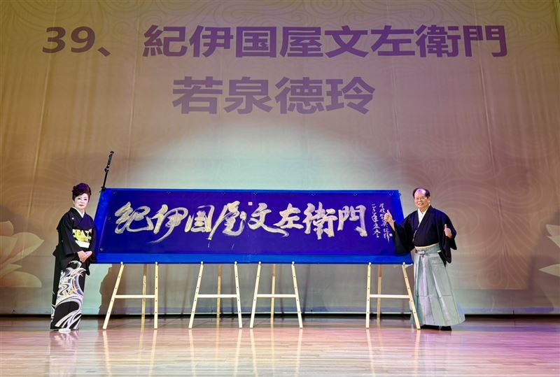 新邦樂舞踊若泉流台灣分家家元若泉德玲（林汶玲老師，左）與林暉博士（右）於舞台上共同展示林博士現場揮毫書法巨作〈紀伊國屋文左衛門〉。作品氣韻生動、筆勢雄健，象徵日台藝術文化的交融與友誼長存。（圖／必紅演藝提供）