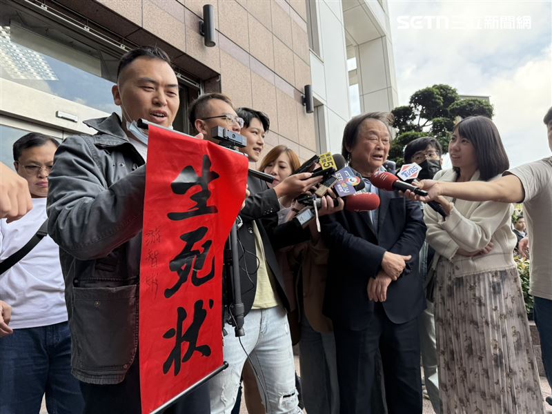 新黨副秘書長游智彬拿著印有「生死狀」的紅布條在王世堅受訪時鬧場（圖／記者劉秀敏攝影）
