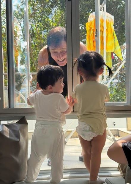 梁家輝身穿黑色背心，站在陽台外隔著玻璃與屋內孫子互動，為了逗笑孩子，梁家輝彎腰貼近窗邊，露出燦爛笑容，甚至笑到「見牙不見眼」。（圖／翻攝自梁靜曦IG）
