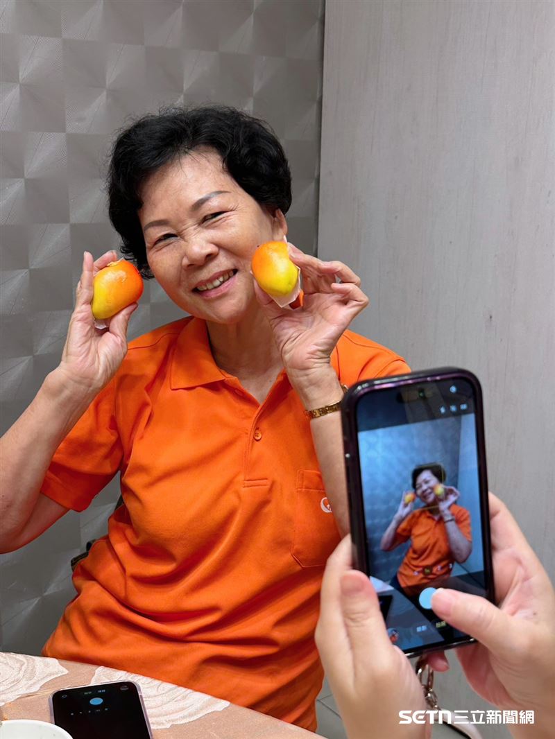 現年73歲的志工林芳芬阿嬤,因拍照姿勢自然又活潑,被大家笑稱是「Pose女王」。面對餐廳特製的芒果造型餐包,她也大方示範拍照,讓現場充滿笑聲。(圖/記者張雅筑攝)