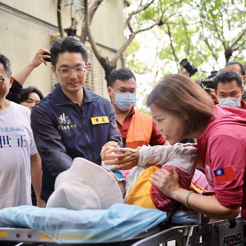 台北市南港區一名參與風災後環境清掃的志工25日身體不適,救護人員協助送上救護車,台北市長蔣萬安(左)在旁握手慰問,關心志工狀況