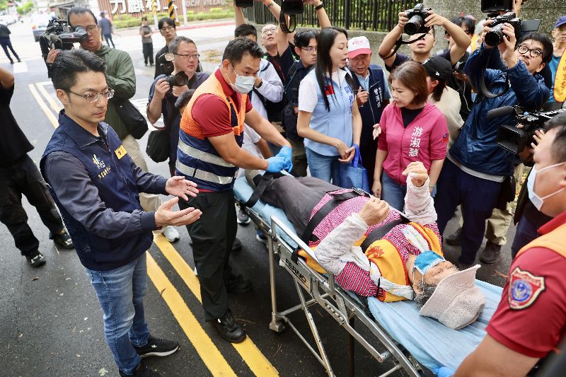 台北市南港區一名參與風災後環境清掃的志工25日身體不適,由救護人員協助躺上擔架,前往巡視復原情形的台北市長蔣萬安(左)跟隨在旁關心